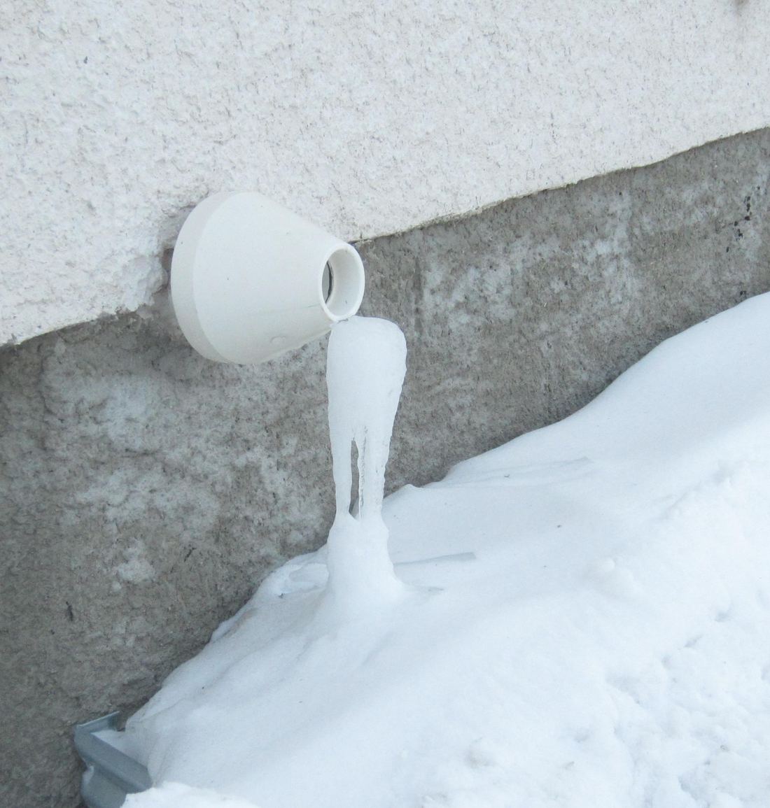 A white plastic vent pipe from a condensing furnace on an outside wall in snow